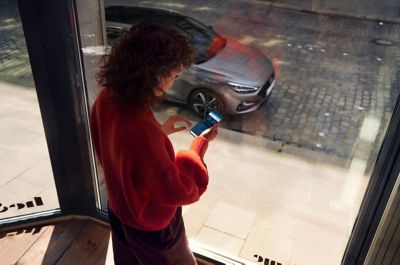 Woman standing by the window, looking at the Bluelink app on her phone. Her Hyundai i30 Fastback is parked on the street. 