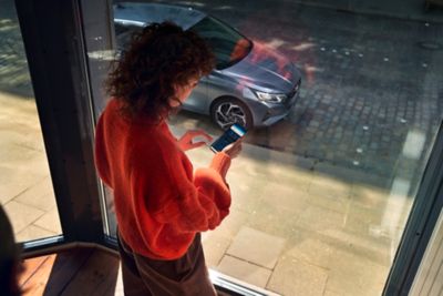 Žena pozerajúca sa na mobilný telefón s aplikáciou Bluelink a nový model i20 zaparkovaný vonku