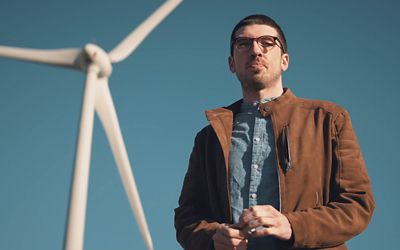 Muž stojaci pred veternou turbínou, ktorá vyrába zelenú elektrickú energiu používanú na nabíjačkách siete IONITY.