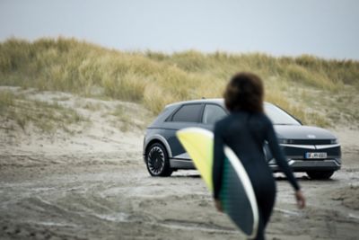 Surferka prichádzajúca k jej elektrickému automobilu Hyundai IONIQ 5 zaparkovanému na pláži.
