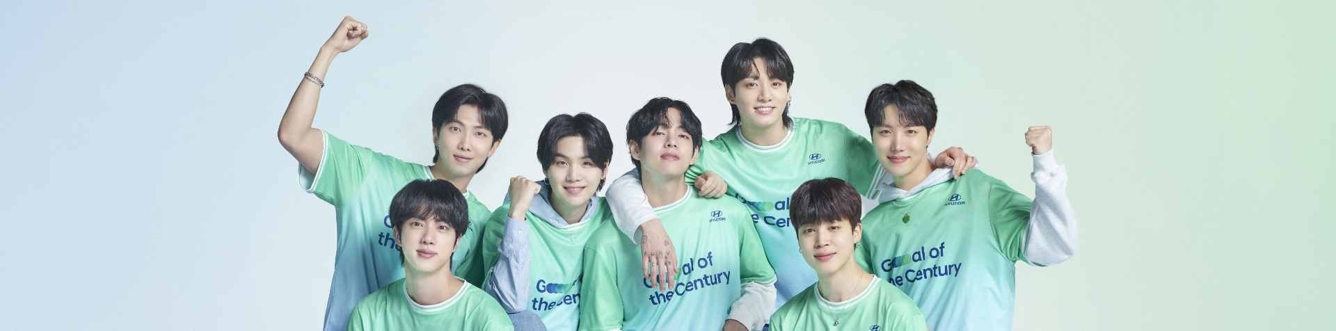 Všetkých 7 členov kapely BTS stojacich v línii smerujúcej dopredu. Každý člen má zelený a sivý dres Team Century s logom Hyundai a tmavomodrým logom Goal of the Century na prednej strane.