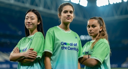 Tri ženy stojace uprostred štadióna v zelených dresoch s nápisom Goal of the Century na prednej strane.